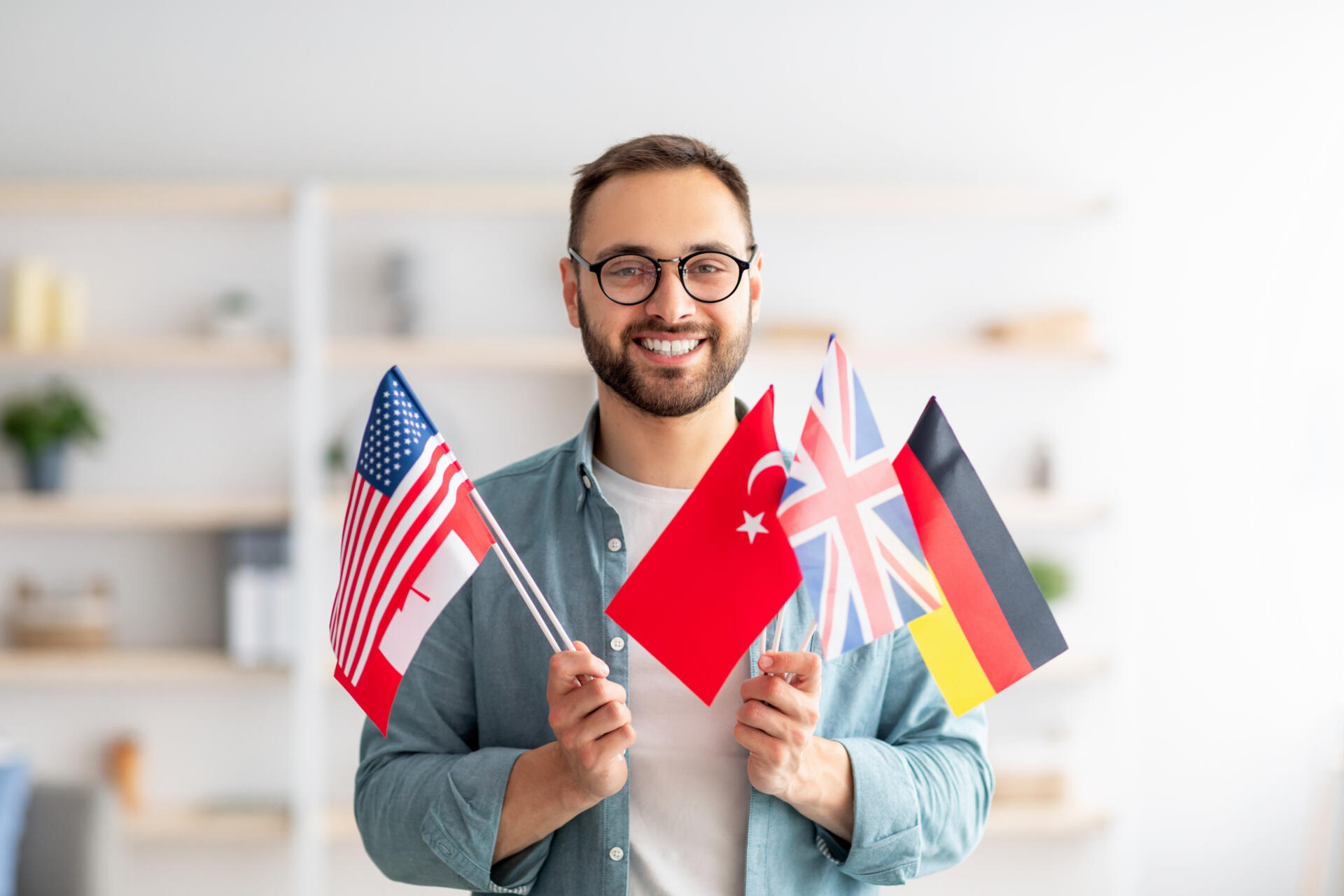 Handsome young guy holding bunch of diverse flags and smiling at camera indoors. Happy millennial man recommending foreign languages school, emigrating abroad. Modern education and student exchange