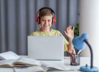 Kako ti gre študij na daljavo? Happy elementary student waving to someone while having an online class over computer at home. Copy space.