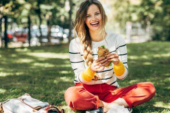 Happy student female sitting on the green grass at the college campus on a sunny day, have lunch and studying outdoors. A smiling young woman takes a rest eating fast food and learning in the park.