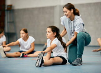 “Veš, Zasavc, svoj šport?” Little girl stretching on the floor and warming up with help of PE teacher during a class at school gym.