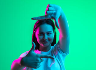 Kakšna je tvoja osebnost? Pleasant emotions. One young beautiful girl demonstrating frame gesture isolated over light green background in neon light. Close-up image