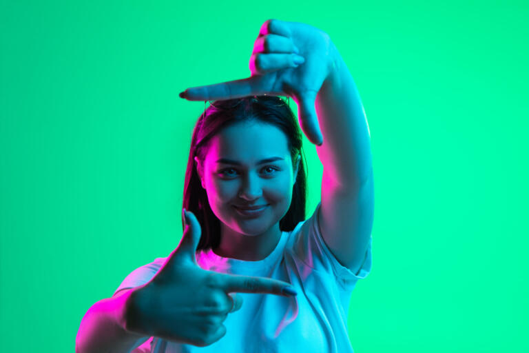 Pleasant emotions. One young beautiful girl demonstrating frame gesture isolated over light green background in neon light. Close-up image