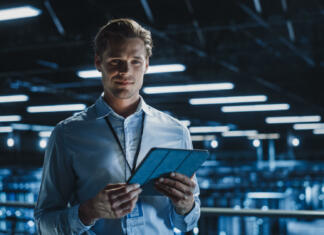 Startup šestnajstletnika vabljen na TechCrunch Disrupt! Portrait of Handsome Smiling IT Specialist Using Tablet Computer in Data Center, Looking at Camera. Succesful Male e-Business Engineer Working in Big Server Farm Cloud Computing Facility.