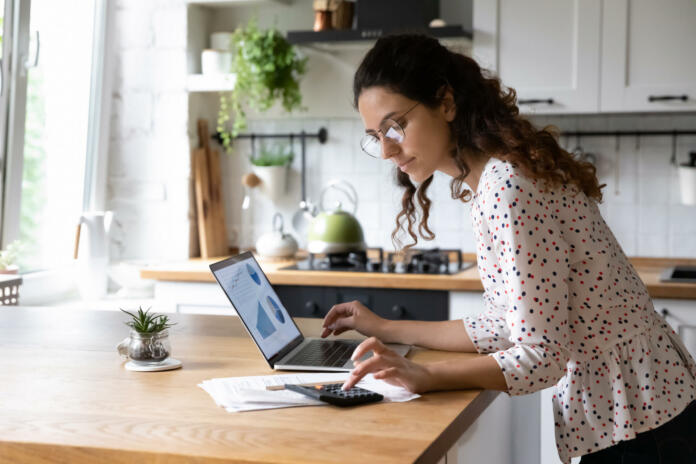Serious young woman wearing glasses calculating finances, household expenses, confident businesswoman working with project statistics, using laptop and calculator, standing in kitchen at home