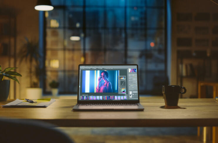 Shot of a Laptop Computer in the Modern Office Showing Photo Editing Software. In the Background Warm Evening Lighting and Open Space Studio with City Window View