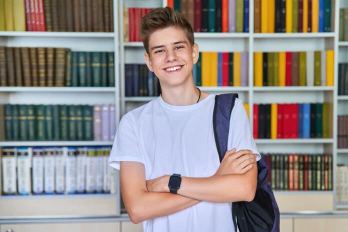 Single portrait of smiling confident male student teenager with backpack with crossed arms looking at camera in the library. School, education, knowledge, adolescence concept