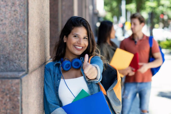 Successful laughing female student showing thumb up with friends in background in front of university building outdoor in summer in city