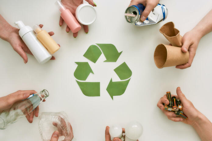 Top view of many hands holding different waste, garbage types with recycling sign made of paper in the center over white background. Sorting, recycling waste concept. Horizontal shot. Top view