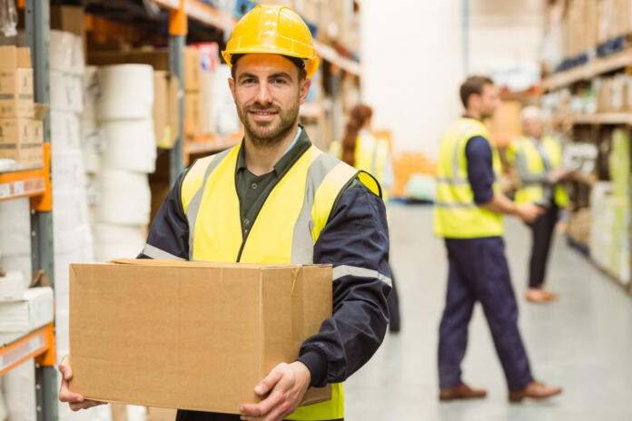 Warehouse worker smiling at camera carrying a box in a large warehouse