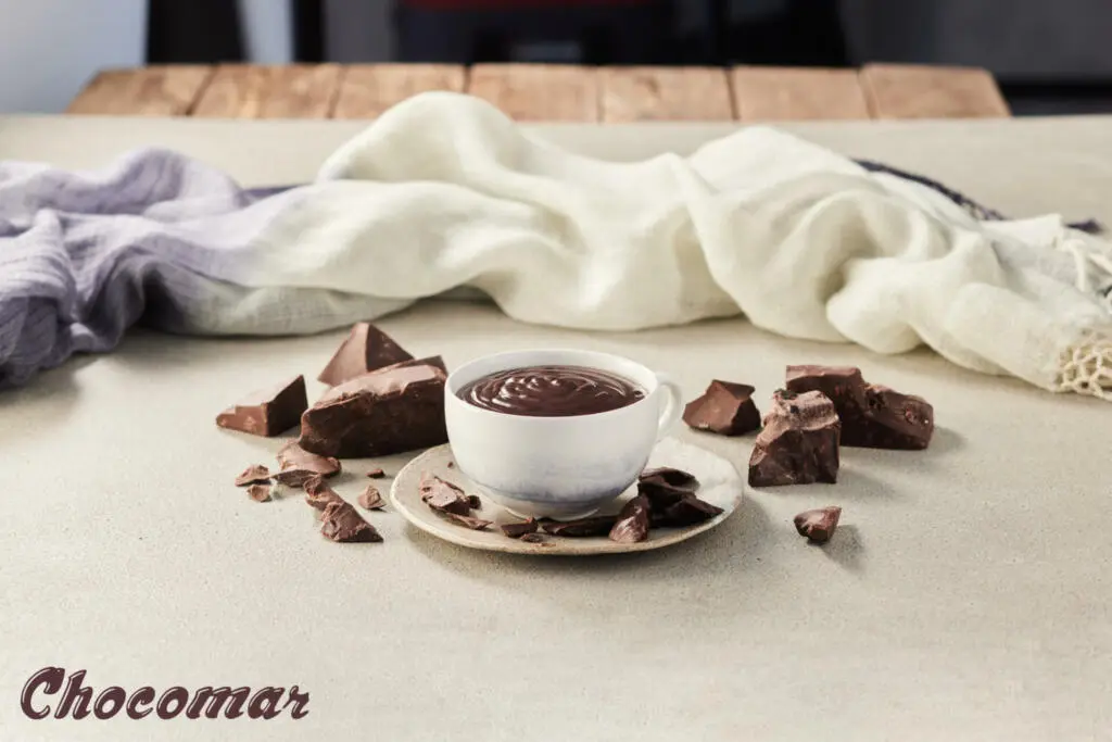 Skodelica vroče čokolade, poleg nje so na mizi koščki čokolade in šal v ozadju