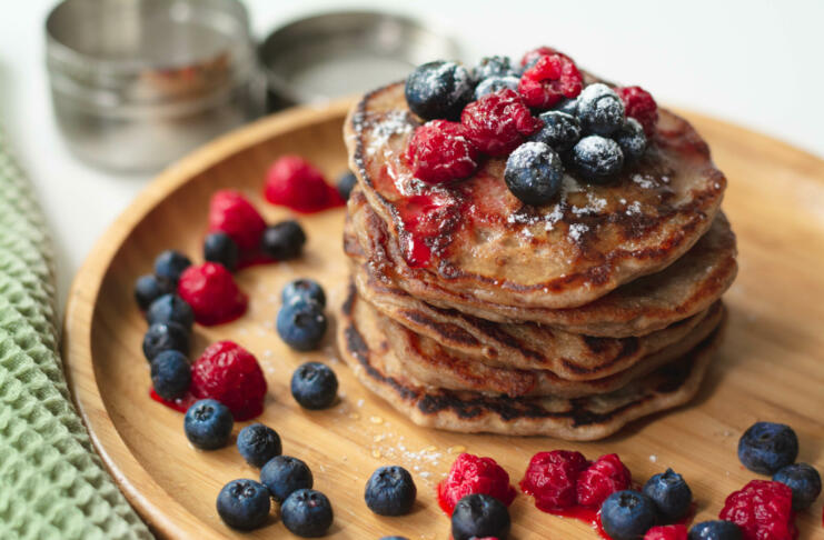 A heap of healthy vegan gluten free whole grain pancakes made with buckwheat flour topped with raspberries and blueberries with icing sugar sprinkled on top