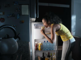 African american woman doing midnight snack at home. She eats a sandwich and looks for food into the refrigerator at night.