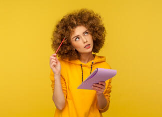 Redno, izredno ali na daljavo? Beautiful pensive woman with Afro hairstyle holding notepad and pencil in hands thinking over plans for startup, wearing casual style hoodie. Indoor studio shot isolated on yellow background.