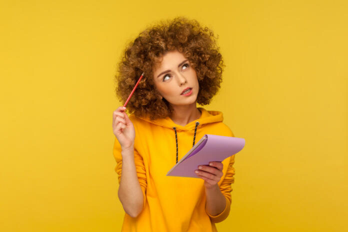 Beautiful pensive woman with Afro hairstyle holding notepad and pencil in hands thinking over plans for startup, wearing casual style hoodie. Indoor studio shot isolated on yellow background.