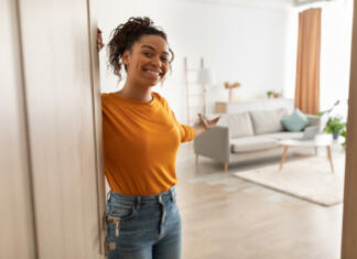 Kako do stanovanja? Cheerful African American Woman Opening Door And Gesturing Welcoming You To Come In Smiling To Camera Standing At Home. Hospitality, Real Estate Ownership And Purchase Concept