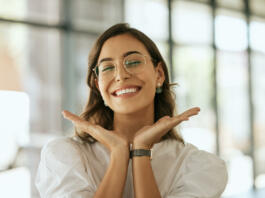 Cheerful business woman with glasses posing with her hands under her face showing her smile in an office. Playful hispanic female entrepreneur looking happy and excited at workplace