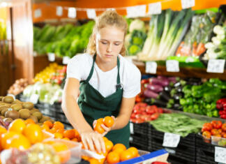Študentsko delo Cheerful young girl employees in uniform holding fresh mandarines in grocery shop