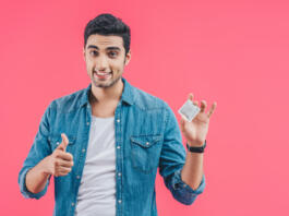 cheerful young man holding condom and doing thumb up gesture isolated on pink
