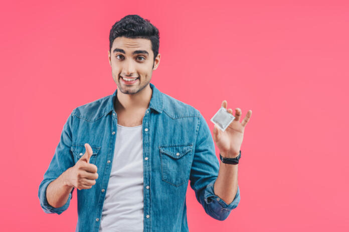 cheerful young man holding condom and doing thumb up gesture isolated on pink