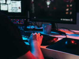 Close up of a gamer montage, detail of the hands of a young man with short blond hair, wearing glasses and headphones, dressed in black shirt, addicted to online video games, playing. Young gamer illuminated with blue and red lights. home office. Dim light from computer monitor, dark room, coloured lights. Horizontal.