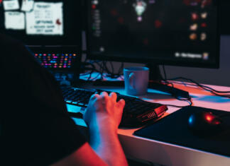 Velik uspeh slovenskih razvijalcev iger Close up of a gamer montage, detail of the hands of a young man with short blond hair, wearing glasses and headphones, dressed in black shirt, addicted to online video games, playing. Young gamer illuminated with blue and red lights. home office. Dim light from computer monitor, dark room, coloured lights. Horizontal.