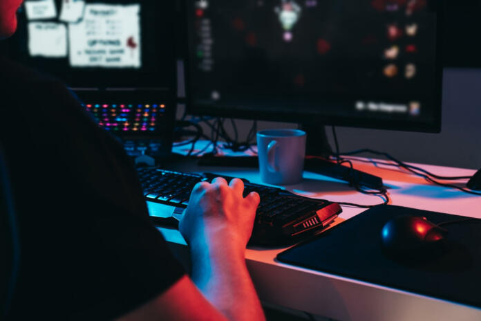 Close up of a gamer montage, detail of the hands of a young man with short blond hair, wearing glasses and headphones, dressed in black shirt, addicted to online video games, playing. Young gamer illuminated with blue and red lights. home office. Dim light from computer monitor, dark room, coloured lights. Horizontal.