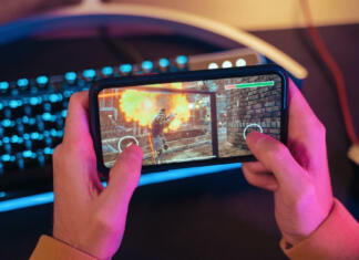 Mobilne igre vodijo v hazarderstvo Close up of male pro gamer's hands holding smartphone with online video shooter game. Cyber sportsman playing mobile games on his cellphone. Professional keyboard on background. Top view. Neon lights