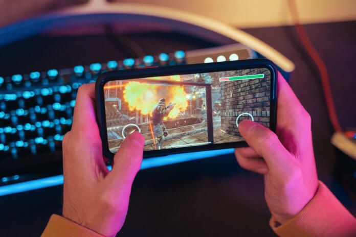 Close up of male pro gamer's hands holding smartphone with online video shooter game. Cyber sportsman playing mobile games on his cellphone. Professional keyboard on background. Top view. Neon lights