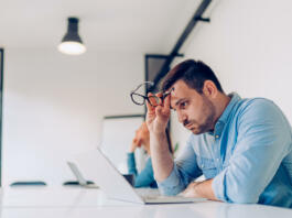 Exhausted young businessman using laptop at work and sitting by the desk while