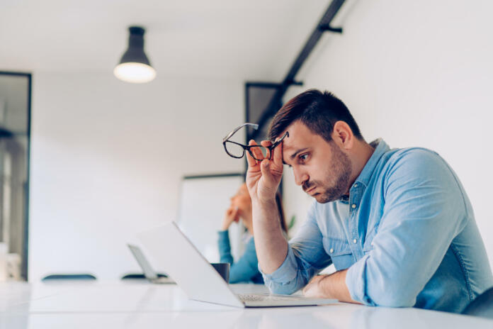 Exhausted young businessman using laptop at work and sitting by the desk while