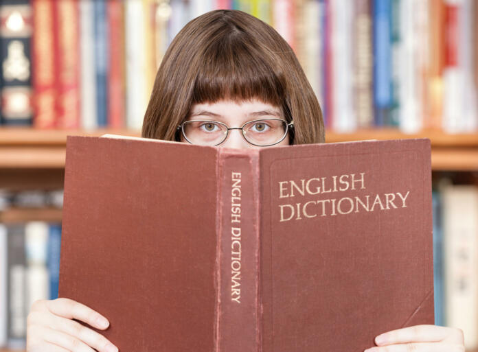 girl with spectacles looks over English Dictionary book and bookcase on background