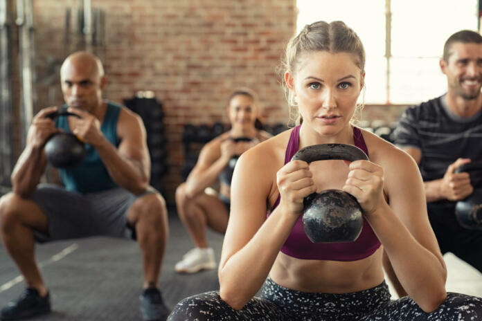 Group of fit people holding kettle bell during squatting exercise at cross training gym. Fitness girl and men lifting kettlebell during strength training exercising. Group of young people doing squat with kettle bell.