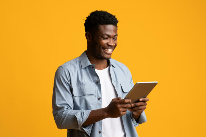 Happy african american millennial guy using modern digital tablet, looking at gadget screen and smiling on yellow studio background, black man gambling or shopping online, copy space