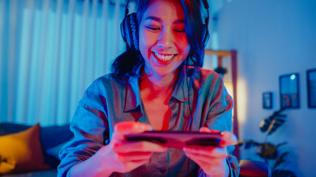 Happy asia girl gamer wear headphone competition play video game online with smartphone colorful neon lights in living room at home. Esport streaming game online, Home quarantine activity concept.