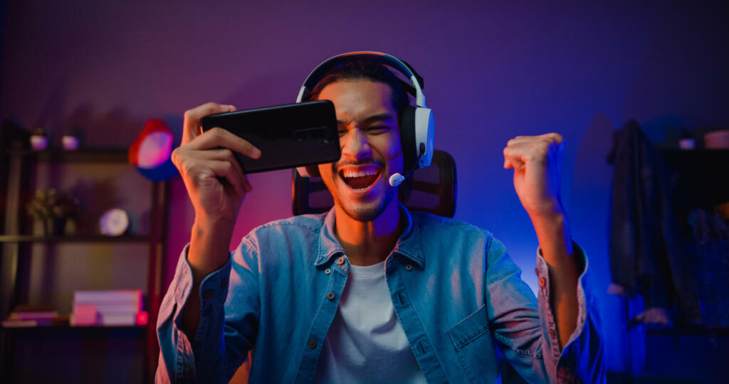 Happy asia man gamer wear headphone competition play video game online with smartphone colorful neon light in living room at night modern house. Esport streaming game online, Home quarantine activity.