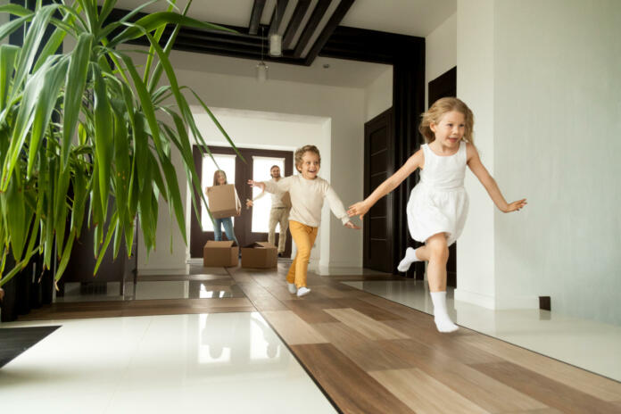 Happy young family with cardboard boxes in new home at moving day concept, excited children running into big modern own house hallway, parents with belongings at background, mortgage loan, relocation