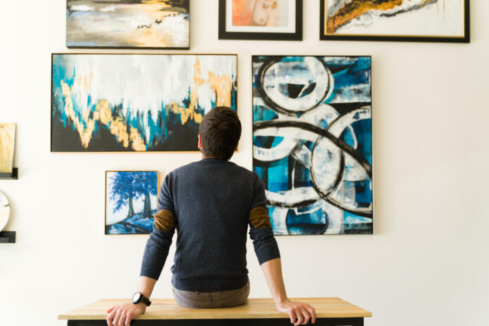 Hispanic male visitor looking reflective while sitting on a bench and admiring the various paintings on the wall of an art gallery