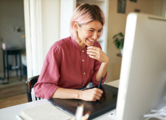 Kakšno je delo urednika portala Študent? Indoor shot of cheerful happy young female with stylish pinkish hair laughing while working from home, sitting at desk with computer and graphic tablet, retouching images or drawing animation