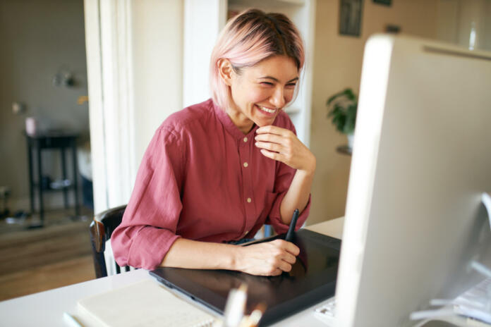 Indoor shot of cheerful happy young female with stylish pinkish hair laughing while working from home, sitting at desk with computer and graphic tablet, retouching images or drawing animation