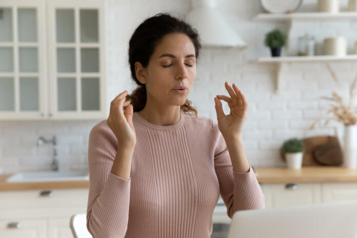 Mindful young woman breathing out with closed eyes, calming down in stressful situation, working on computer in modern kitchen. Millennial hispanic lady managing stress, practice yoga at home office.