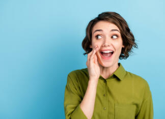 Ženske lažje najdejo prave besede? Photo portrait of young pretty girl with short hair, telling secret information rumouring gossiping wearing green shirt isolated on blue color background