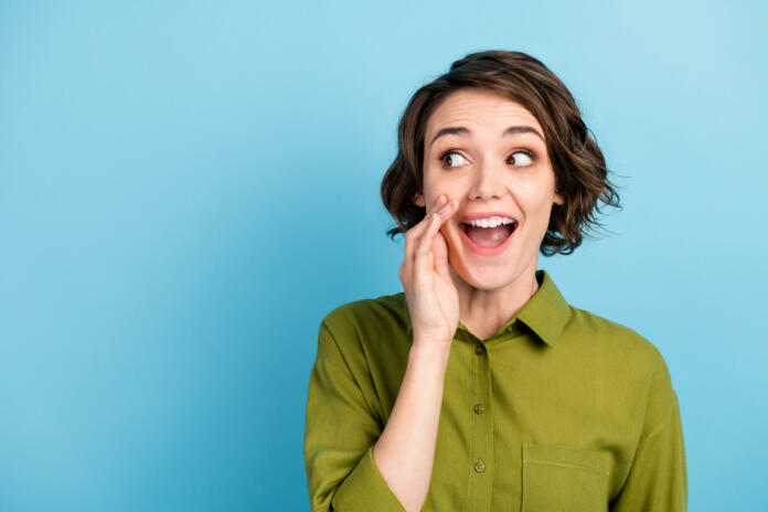 Photo portrait of young pretty girl with short hair, telling secret information rumouring gossiping wearing green shirt isolated on blue color background