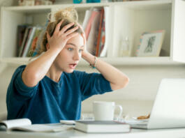 Portrait of an attractive woman at the table with cup and laptop, book, notebook on it, grabbing her head. Bookshelf at the background, concept photo