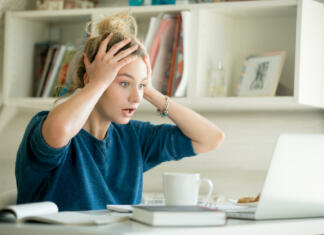 Nisi sprejet? Ni panike Portrait of an attractive woman at the table with cup and laptop, book, notebook on it, grabbing her head. Bookshelf at the background, concept photo
