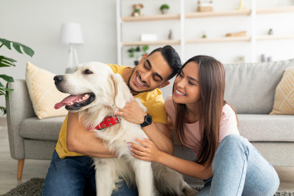 Portrait of happy multiracial couple scratching their pet dog, sitting on floor at home. Arab guy and his Caucasian girlfriend hugging their golden retriever in living room