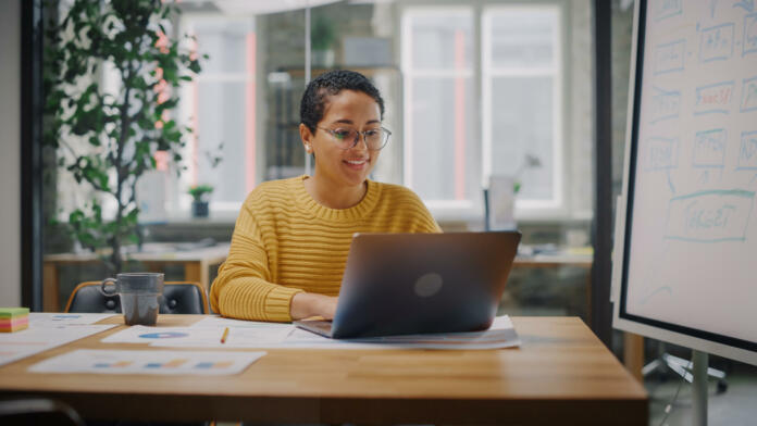 Portrait of Young Latin Marketing Specialist in Glasses Working on Laptop Computer in Busy Creative Office Environment. Beautiful Diverse Multiethnic Female Project Manager is Browsing Internet.