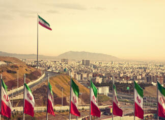 Presenetljiva dežela Perzijcev Set of Iran flags in Front of Tehran Skyline and one large flag in the background at sunset with orange warm tone.