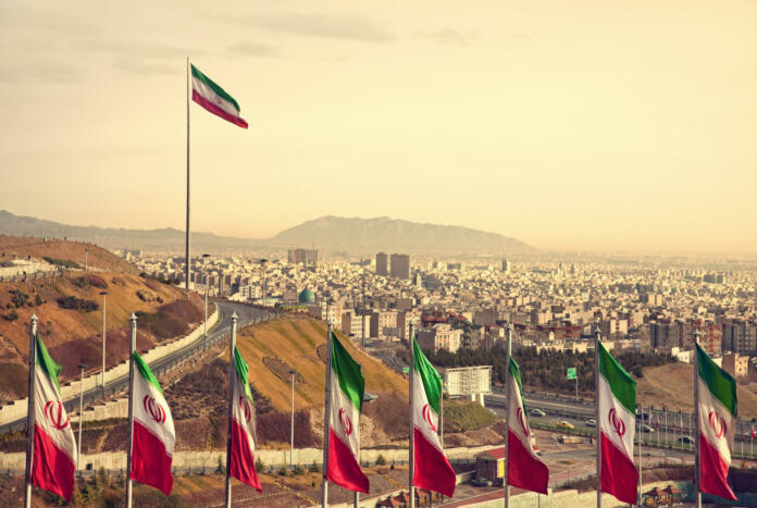 Set of Iran flags in Front of Tehran Skyline and one large flag in the background at sunset with orange warm tone.