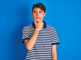 Teenager boy wearing casual t-shirt standing over blue isolated background Thinking worried about a question, concerned and nervous with hand on chin