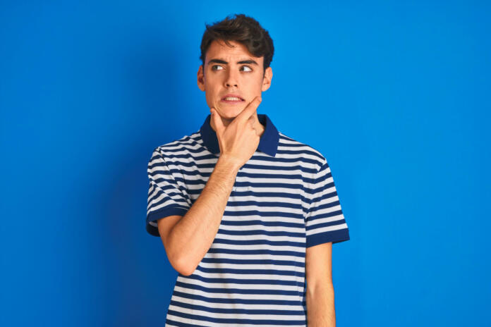 Teenager boy wearing casual t-shirt standing over blue isolated background Thinking worried about a question, concerned and nervous with hand on chin